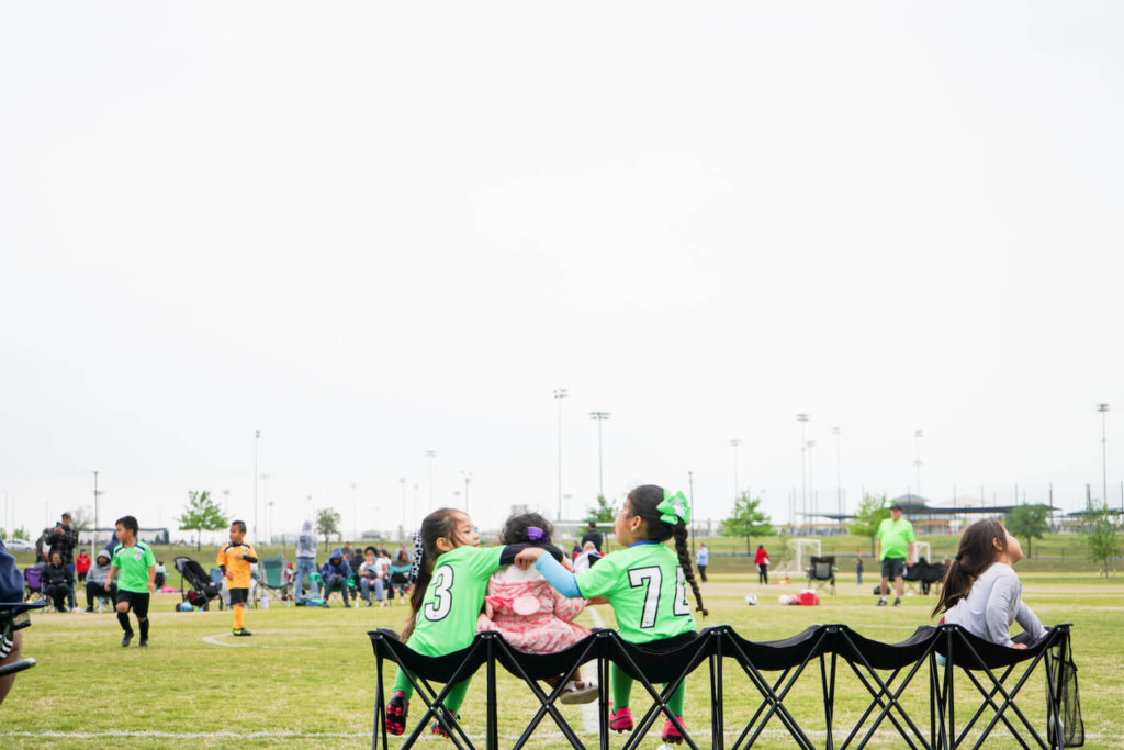 Girls on a soccer field sitting on the sidelines playing with each other.